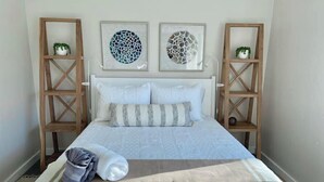 Ein ordentlich bezogenes Bett mit weißer Steppdecke und Kissen, zwei Wandregale mit Pflanzen und zwei gerahmte abstrakte Kunstwerke.