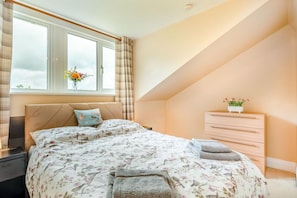 Un dormitorio con cama, cómoda, ventana con cortinas y un florero con flores.