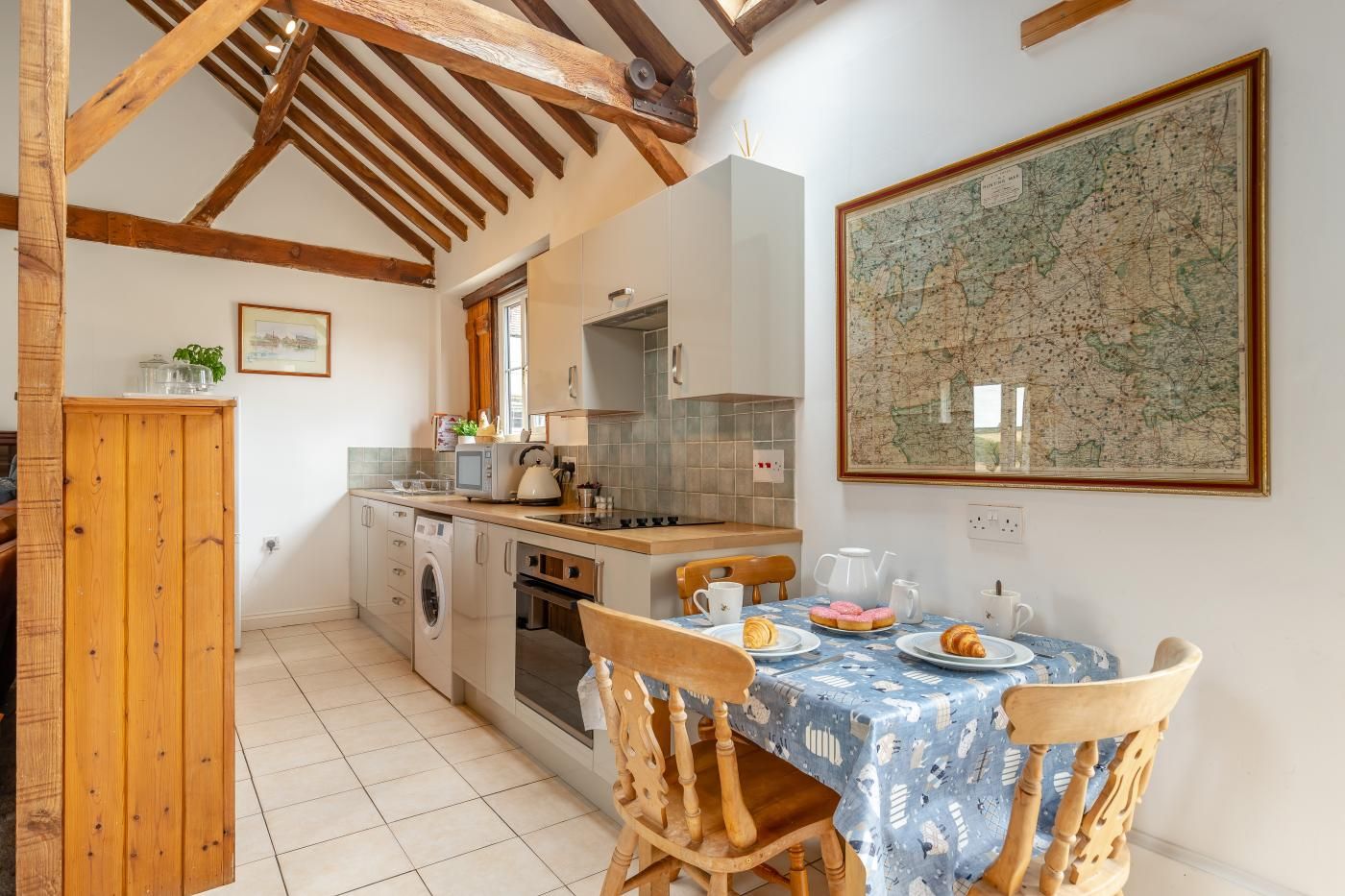 Une cuisine avec des poutres en bois, une table à manger recouverte d’une nappe bleue et une carte accrochée au mur.
