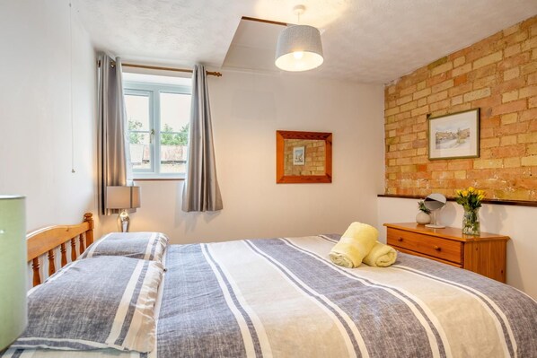 A bedroom with a bed, a window with curtains, a brick wall, and a wooden dresser with a mirror and vase.