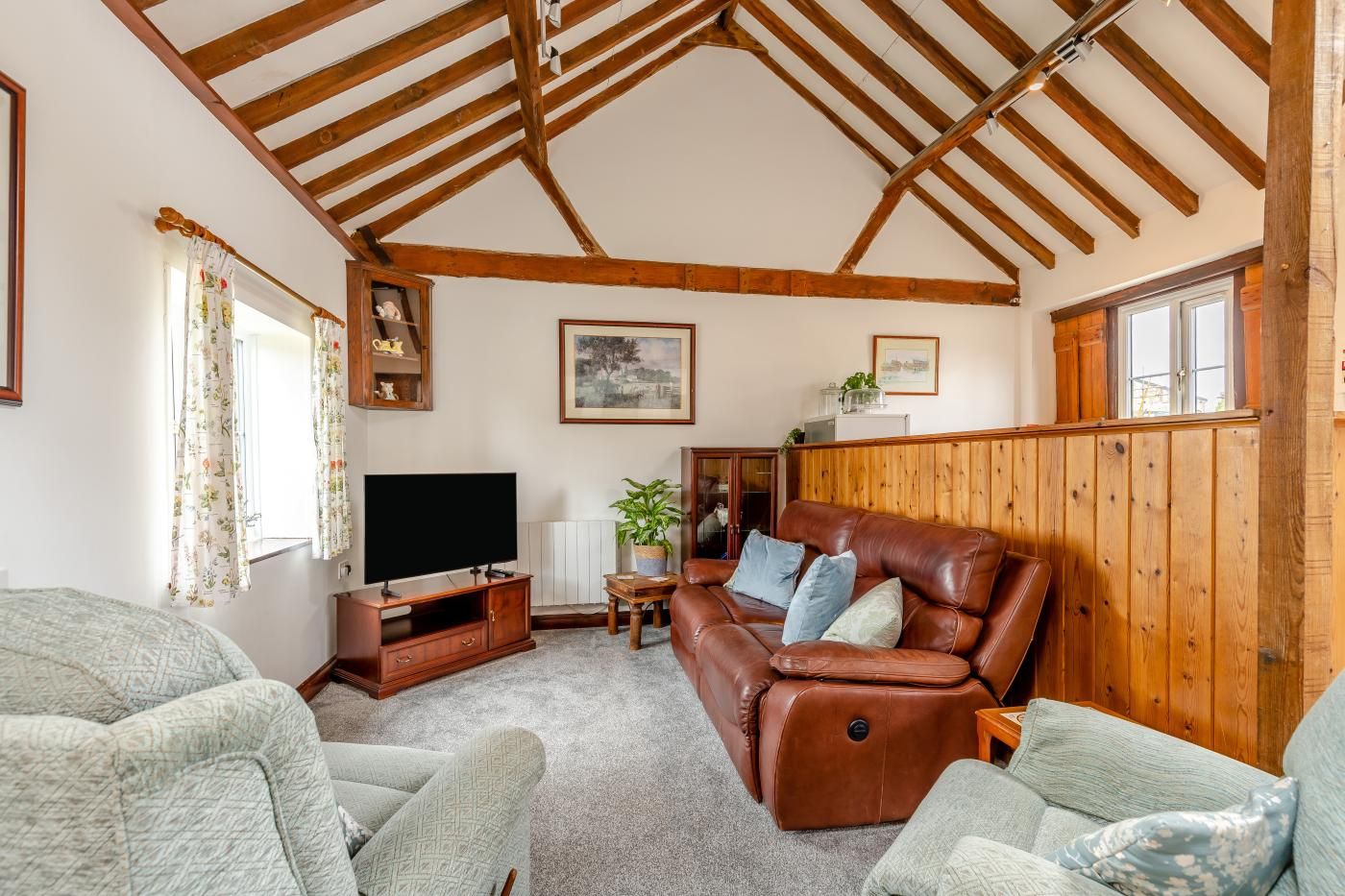 A cozy living room with a wooden ceiling, a flat-screen TV, a brown leather sofa, and a patterned armchair.