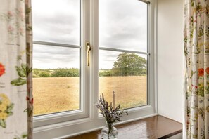 Una ventana con vista a un campo y árboles, un florero con flores en el alféizar, y cortinas con motivos florales.