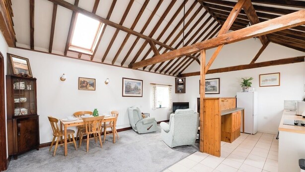 A spacious room with a high ceiling, wooden beams, a dining area with chairs and a table, a sofa, a small kitchen area with a fridge, and a window.