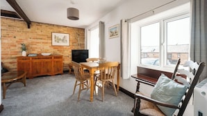 A cozy living room with a brick wall, wooden furniture, a TV, and a large window. - 2 Bed in Thurleigh (oc-o42689) (Thurleigh)