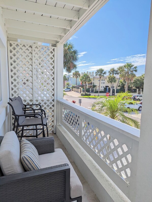 Un balcon doté d’une balustrade blanche en treillis, de mobilier d’extérieur et offrant une vue sur une rue résidentielle bordée de palmiers et de voitures garées.