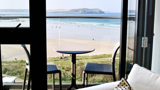 Habitación con un amplio ventanal que da a una playa, una mesa redonda, dos sillas y vistas al océano y colinas lejanas.