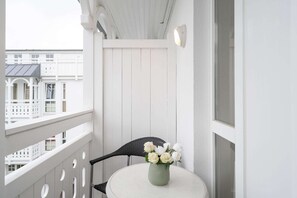 Un balcón con barandilla blanca, una mesa redonda, un jarrón verde con flores blancas y una silla negra.