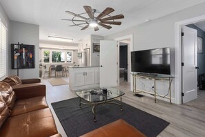 A modern living room with leather sofas, a glass coffee table, a ceiling fan, a TV on a stand, and a kitchen in the background. - Single Level Ranch - 2 Bedroom Home - sleeps 4 (North Kansas City)