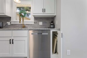 A kitchen with white cabinets, a stainless steel dishwasher, and a window with a view of trees. - Single Level Ranch - 2 Bedroom Home - sleeps 4 (North Kansas City)