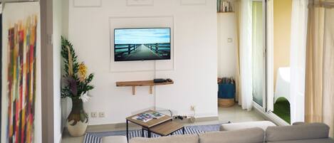 A modern living room with a large flat-screen TV, a sofa, a coffee table, a ceiling fan, and a view through a sliding glass door.