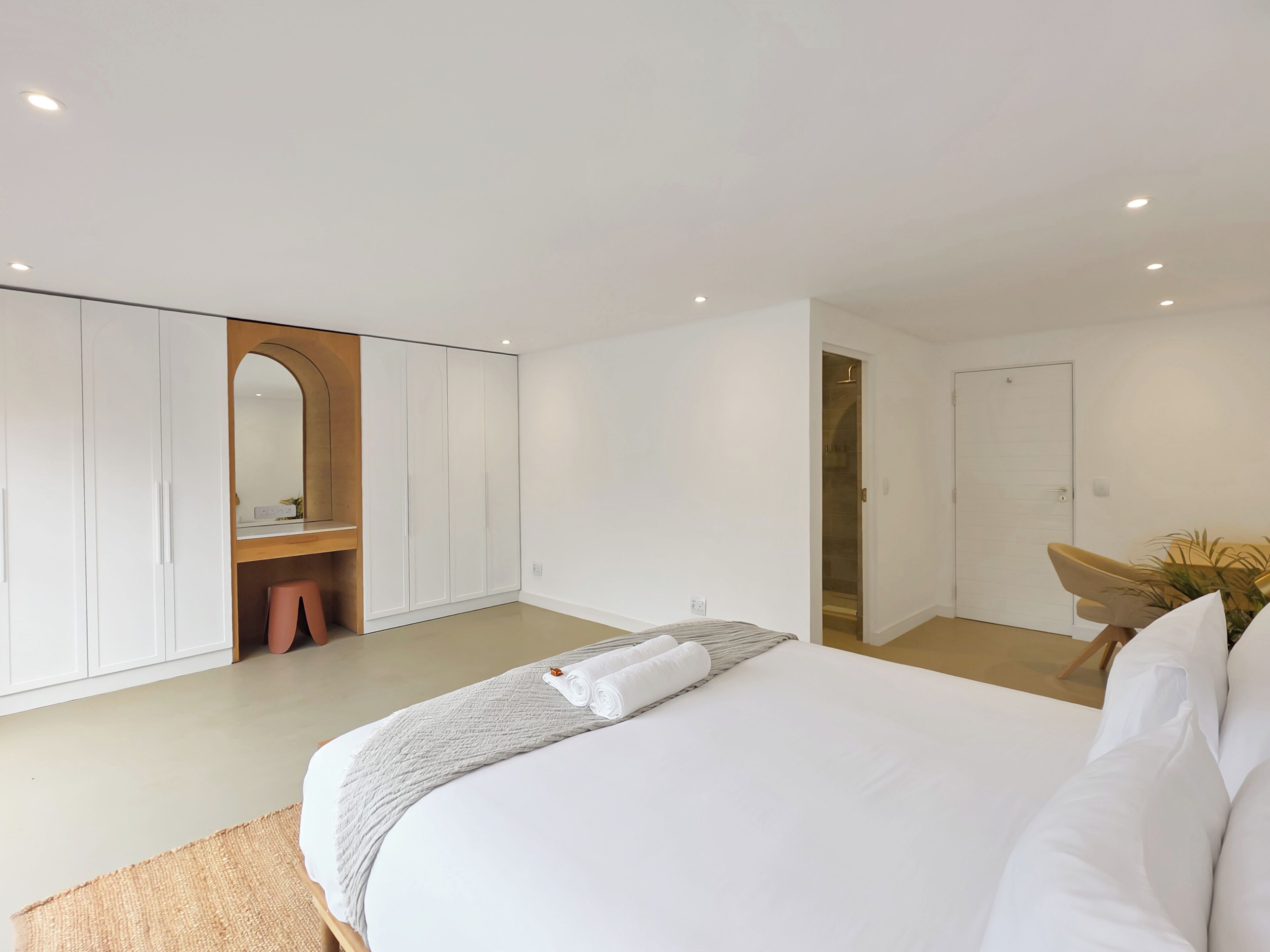 A modern hotel room with a large bed, a wardrobe, a vanity with a mirror, and a door leading to another room.