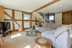 Un salon avec des poutres apparentes, un canapé, une table basse en verre, un téléviseur et un escalier.