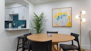 Uma cozinha moderna com armários brancos e painel de parede azul, uma área de jantar com mesa de madeira redonda e cadeiras pretas, um grande quadro abstrato na parede e um abajur de chão com várias luzes.