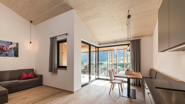A modern living room with a wooden ceiling, a dining area with a table and chairs, a sofa with red cushions, and a kitchen area with a stove. - Cozy vacation rental with a spacious loggia and a magnificent view (Trentino-Alto Adige)