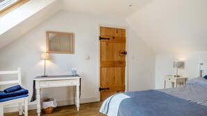A bedroom with a sloped ceiling, a wooden door, a bedside table with a lamp, a bed with a striped blanket, and a chair with blue cushions. - 2 Bed in Dwrbach (oc-w42727) (Dwrbach)
