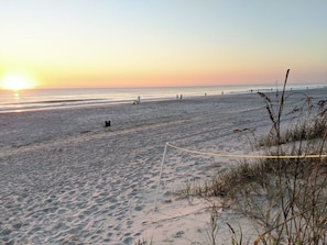 Ein Strand mit Volleyballnetz, Spaziergängern und Sonnenuntergang.