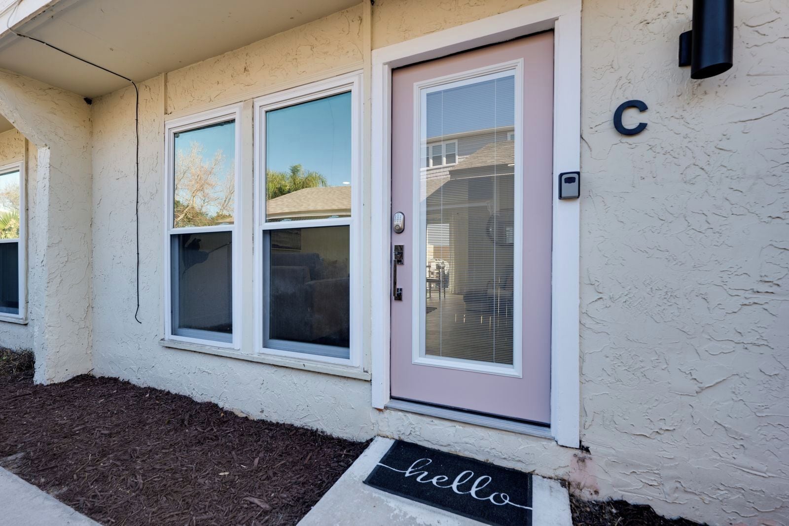 Une maison avec une porte d’entrée, deux fenêtres, un paillasson où est écrit « Bonjour » et un panneau en forme de « C » sur le mur.