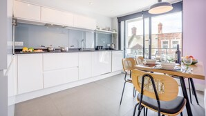 A modern kitchen with white cabinets, a dining area with wooden chairs, and a large window with a city view. - 2 Bed in Swanage (oc-c41747) (Swanage)