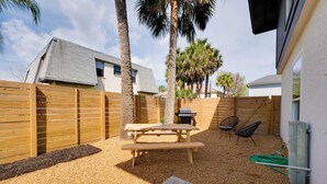 Un jardin avec une table de pique-nique, deux chaises, un barbecue et une clôture en bois.