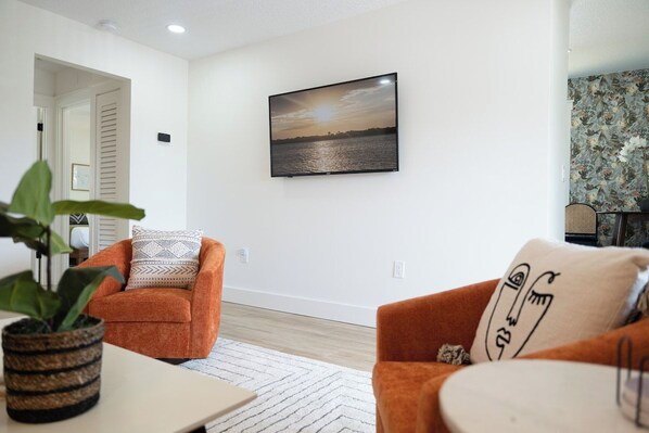 A living room with a flat-screen TV mounted on the wall, a sofa with patterned cushions, a potted plant, and a view of a sunset over water. - Be A Nomad | New Listing l 2br 3 blks to beach - D (Jacksonville Beach)
