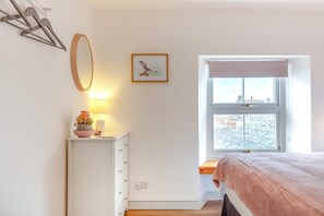 A bedroom with a bed, a bedside table with a lamp, a window with a view of buildings, and a wall-mounted mirror. - 2 Bed in Lynton (oc-v42649) (Lynmouth)