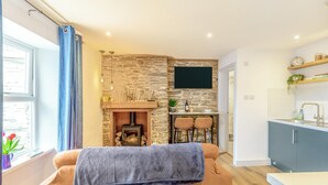 A cozy living room with a fireplace, a TV mounted on a stone wall, a dining area with two chairs, and a kitchen with a sink and shelves. - 2 Bed in Lynton (oc-v42649) (Lynmouth)