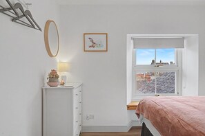 Ein Schlafzimmer mit einem Bett, einem Nachttisch mit Lampe, einem Fenster mit Blick auf Gebäude und einem an der Wand befestigten Spiegel.