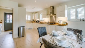 A modern kitchen with white cabinets, a dining area with a table set for four, and a view of the outside through a window. - 4 Bed in Mundesley (oc-s42724) (Mundesley)