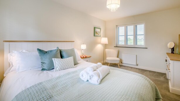 A bedroom with a bed, pillows, a bedside table, a chair, a window, a radiator, and a chandelier. - 4 Bed in Mundesley (oc-s42724) (Mundesley)