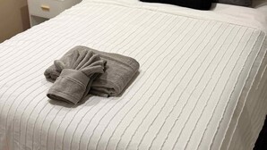 Un lit bien fait, recouvert d’une couverture rayée blanche, avec une serviette grise pliée et deux oreillers, un gris et un noir, dans une pièce où un tableau représentant un pont est accroché au mur.