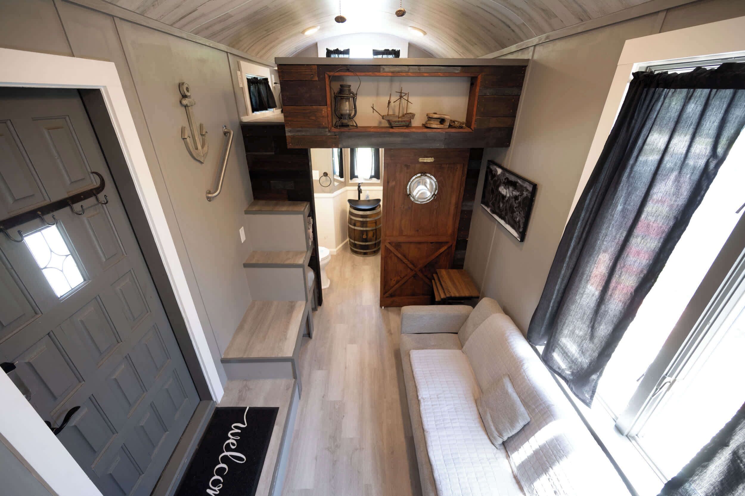 Un intérieur compact comprenant un canapé, une étagère en bois agrémentée d’objets décoratifs, une porte avec une poignée et une fenêtre avec des rideaux.