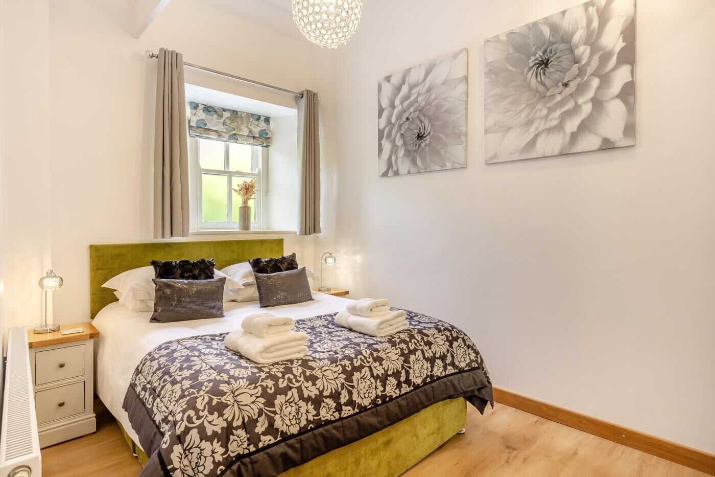 Une chambre à coucher comprenant un lit, une table de chevet, une lampe, une fenêtre avec des rideaux et deux tableaux représentant des fleurs, accrochés au mur.