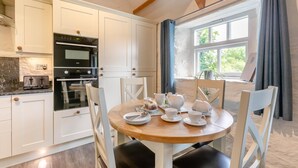 A kitchen with white cabinets, a wooden dining table set with tea cups, and a view of greenery outside. - 2 Bed in Duloe (oc-r27525) (Duloe)