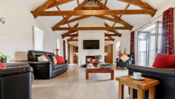 A spacious living room with a high ceiling, wooden beams, a fireplace, and a television. - 3 Bed in Duloe (oc-h27526) (Duloe)