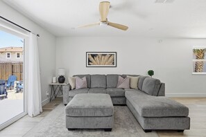 Ein Wohnzimmer mit grauem Ecksofa, Deckenventilator, gerahmtem Kunstwerk und Blick ins Freie.