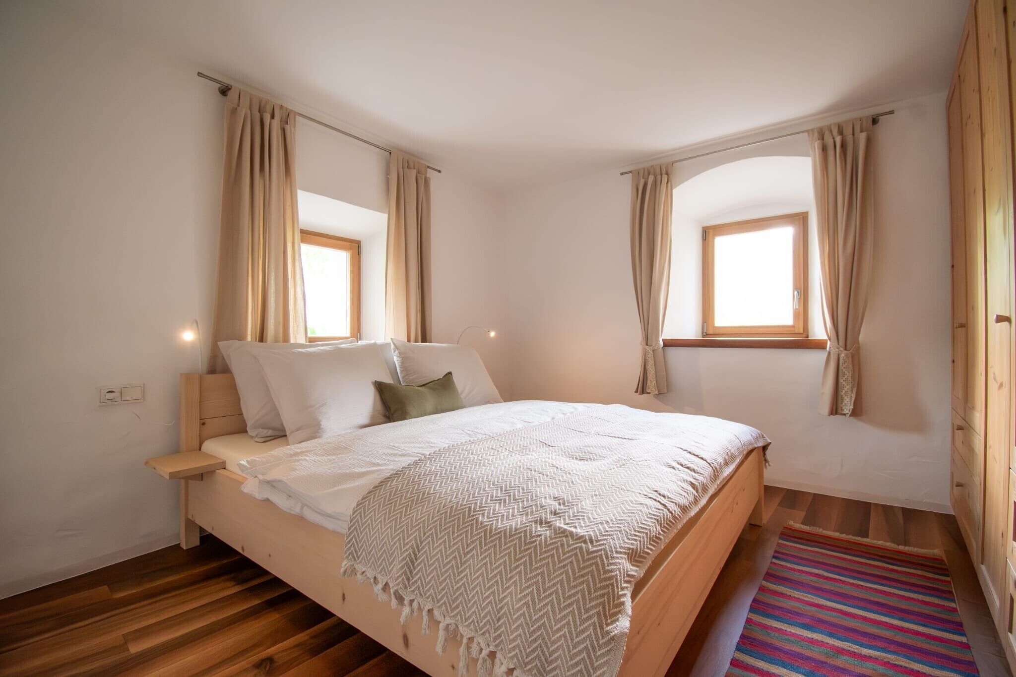 A bedroom with a wooden bed, two windows with curtains, a bedside table, and a colorful rug.