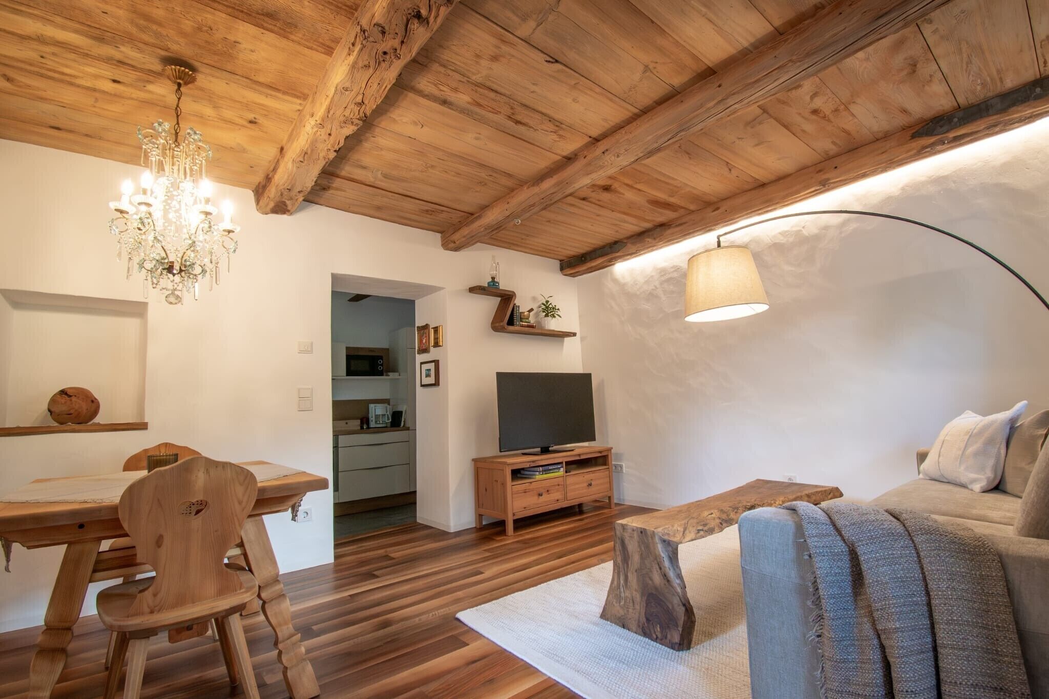 A wooden ceiling, a chandelier, a television on a wooden stand, a sofa, and a dining table with chairs.