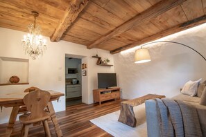 Techo de madera, una araña, un televisor sobre un mueble de madera, un sofá y una mesa de comedor con sillas.