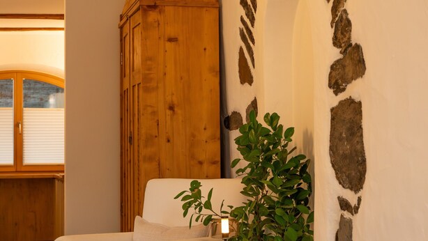 A room with a white armchair, a wooden cabinet, a potted plant, and a stone archway on the wall. - Saxöler – Cozy Apartment in Historic Winery near Brixen (Brixen)