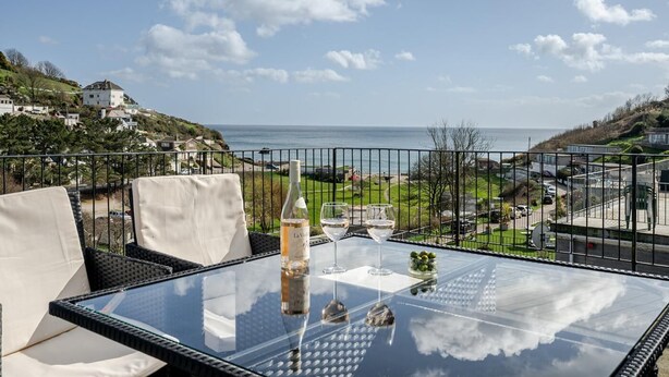 Une table à plateau en verre, avec une bouteille et des verres, donnant sur un paysage côtier avec des maisons et de la végétation.