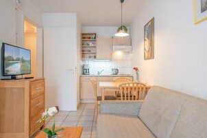 Un espacio habitable compacto con kitchenette, comedor, sofá, televisor y una planta.
