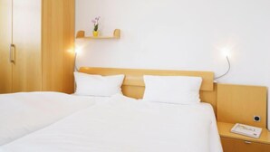 Quarto de hotel com cabeceira de madeira, duas camas brancas, mesa de cabeceira com abajur e uma pequena prateleira com uma flor.
