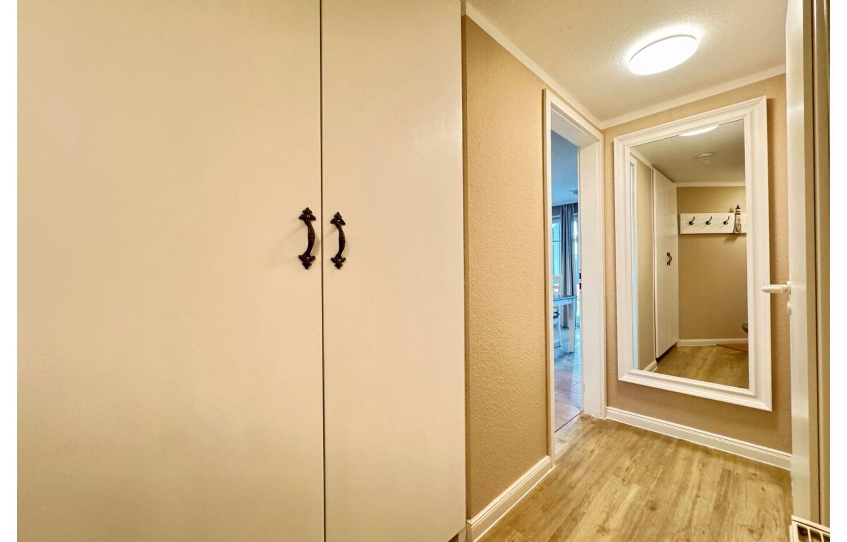 Un couloir doté d’un plancher en bois, de murs de couleur beige et d’un miroir qui reflète une pièce où se trouve un porte-manteau.