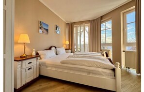 A hotel room with a large bed, two bedside tables, a window with curtains, and a view of the outdoors. - Urlaub auf Rugen mit Ahoi-vorteil (Sellin)