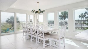 A spacious dining area with a white table and chairs, large windows, and a modern chandelier. - Be a Nomad | Stunning Oceanfront 7-bedroom Home (Jacksonville Beach)
