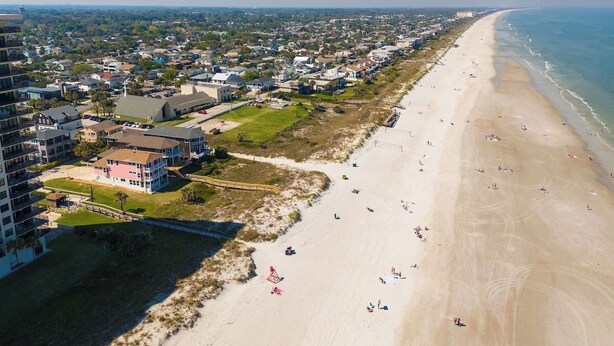 An aerial view of a coastal area with a beach, residential buildings, and a clear sky. - Be a Nomad | Stunning Oceanfront 7-bedroom Home (Jacksonville Beach)