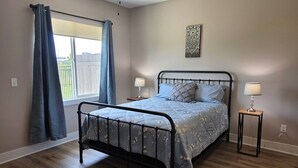 A bedroom with a metal bed frame, a window with curtains, a bedside table with a lamp, and a patterned rug. - New Construction Zero Entry in N Wichita (Wichita)