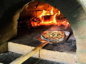 A pizza oven with a pizza on a metal tray, a wooden pizza peel, and a fire burning in the background.
