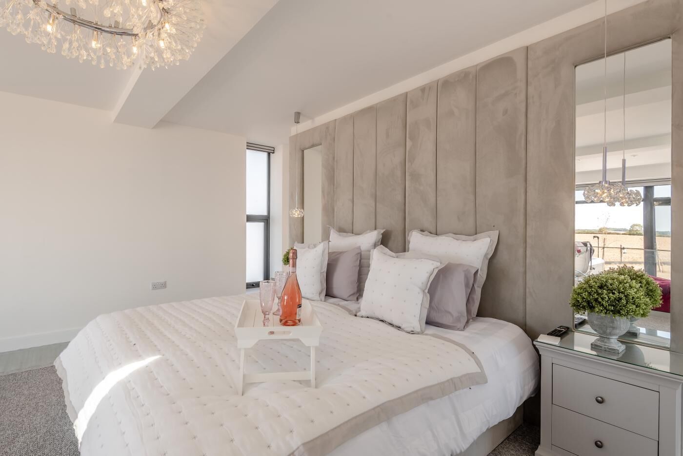A modern bedroom with a large bed, a bedside table, a mirror, and a chandelier.