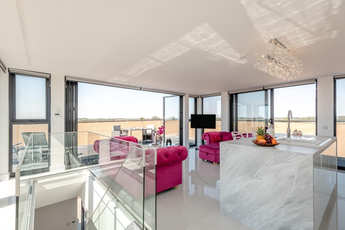 A modern living room with a glass railing, a pink sofa, a marble island, and a view of the desert.